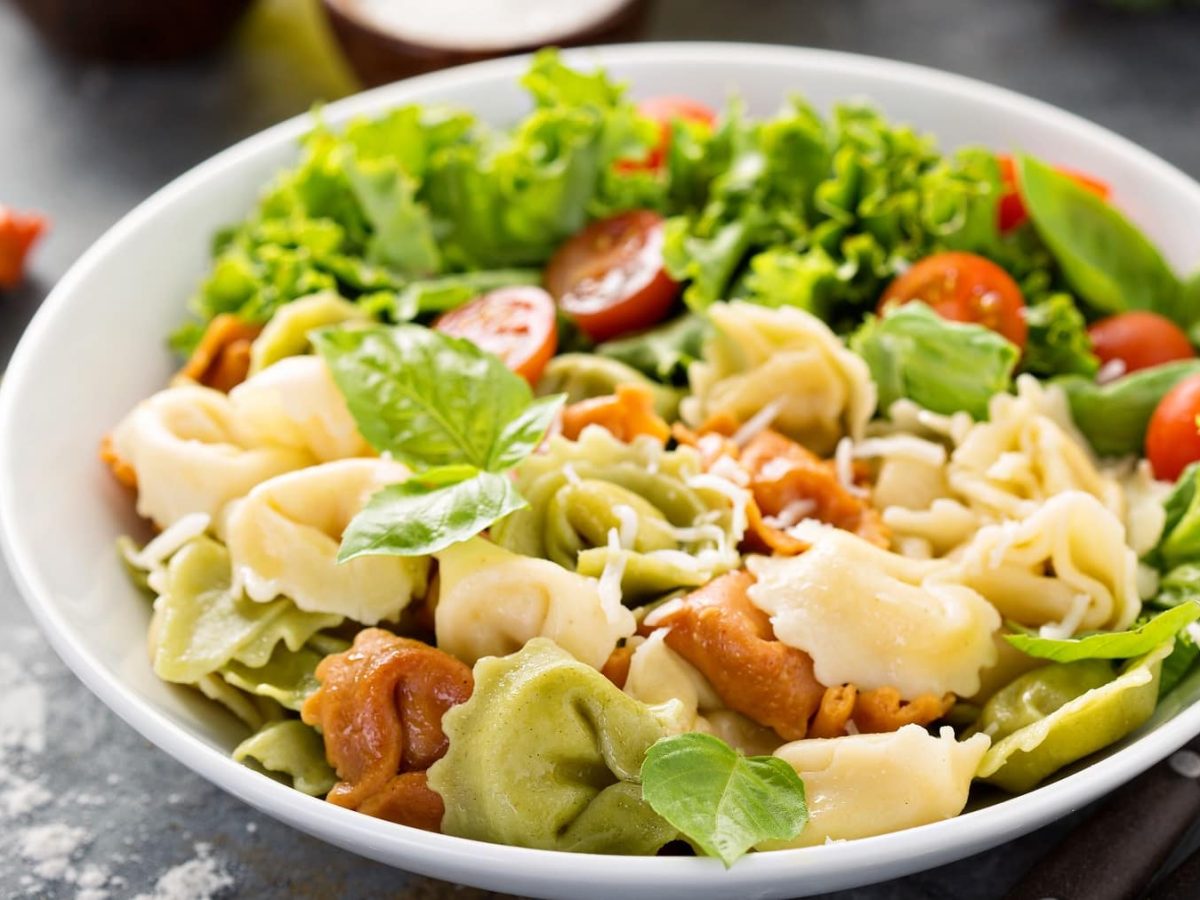 Ein weißer Teller mit dem Tortellini-Salat auf einem grauen Steintisch. Daneben eine Schale mit dem Dressing und einzelne Tortellini.