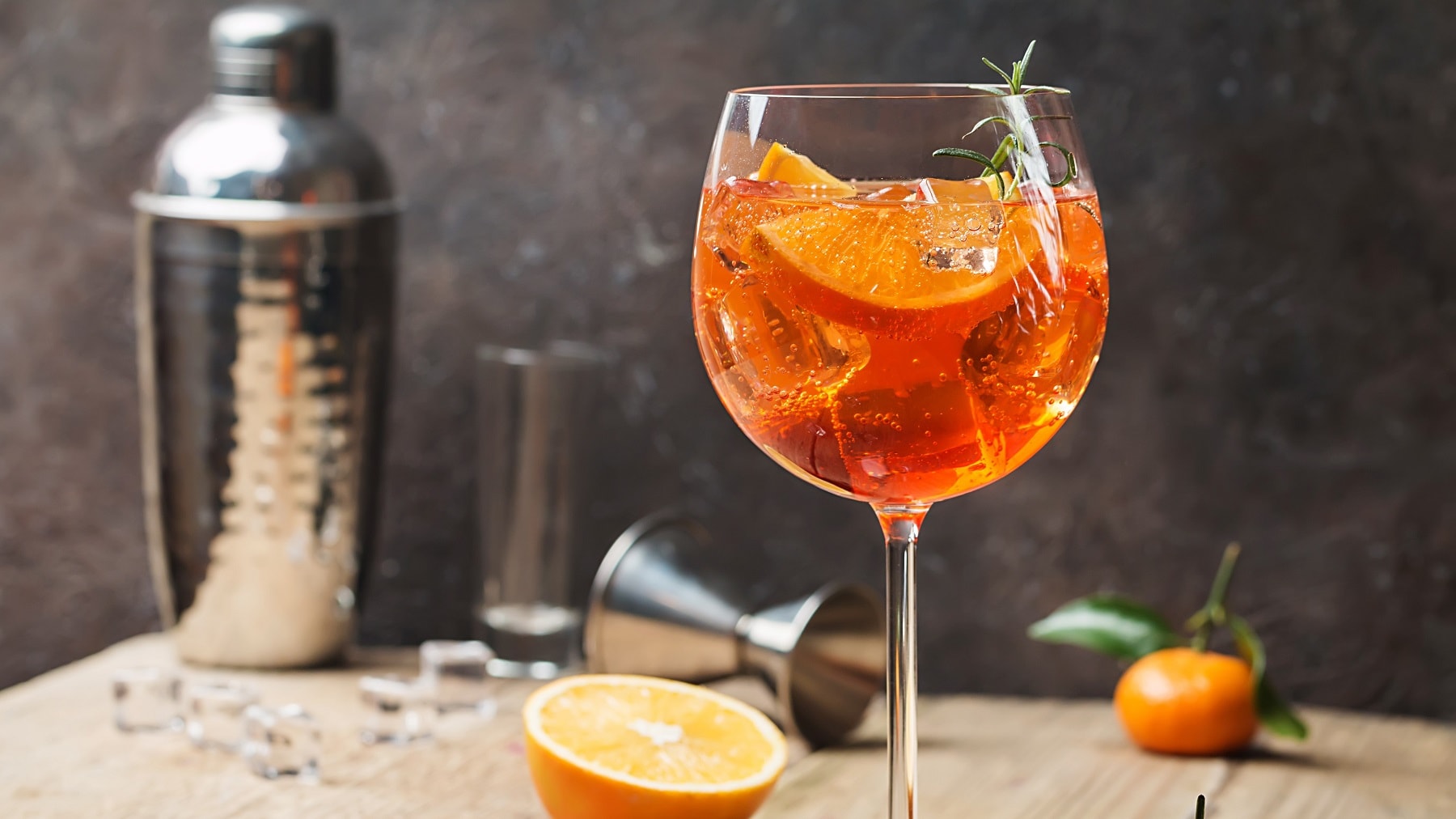 Ein Glas mit dem Canonita-Spritz, vielen Orangen und Cocktailzubehör auf einem Holztisch und grauer Wand.