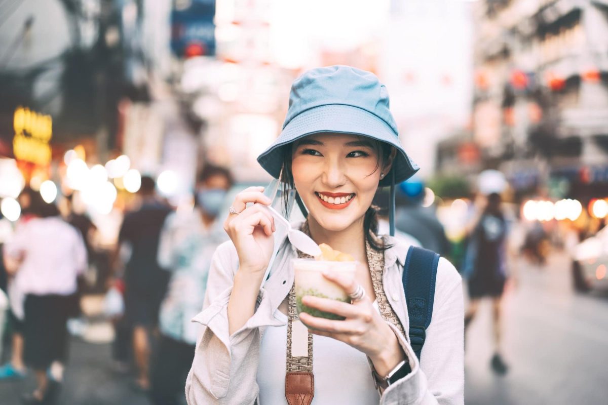 Vegane Städte: asiatische Touristin mit Hut, Rucksack und Eiscreme in der Hand, lächelnd, im Hintergrund unscharf eine Stadt.