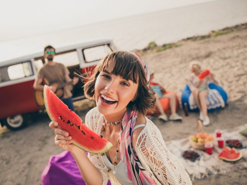 Frontal: Junge Menschen picknicken am Strand. Im Vordergrund ist eine junge Frau, die in eine Melonen-Scheibe in der Hand hält. Im Hintergrund ist ein VW-Bus.
