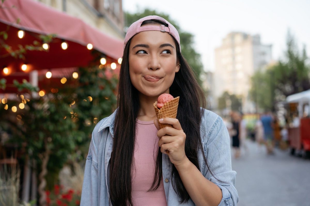 Eis-Umfage Hellofresh: Eine Frau mit dunklen, lagen Haaren und Basecap hält eine Waffel mit Vanilleeis in der Hand, im Hintergrund unscharf eine Ladenmarkise.