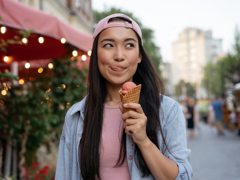Eis-Umfage Hellofresh: Eine Frau mit dunklen, lagen Haaren und Basecap hält eine Waffel mit Vanilleeis in der Hand, im Hintergrund unscharf eine Ladenmarkise.