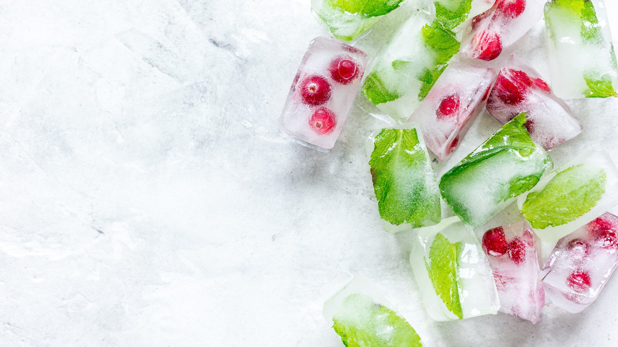 Draufsicht: Auf einer weißen Oberfläche aus Eis liegen Früchte-Eiswürfel und Kräuter-Eiswürfel.