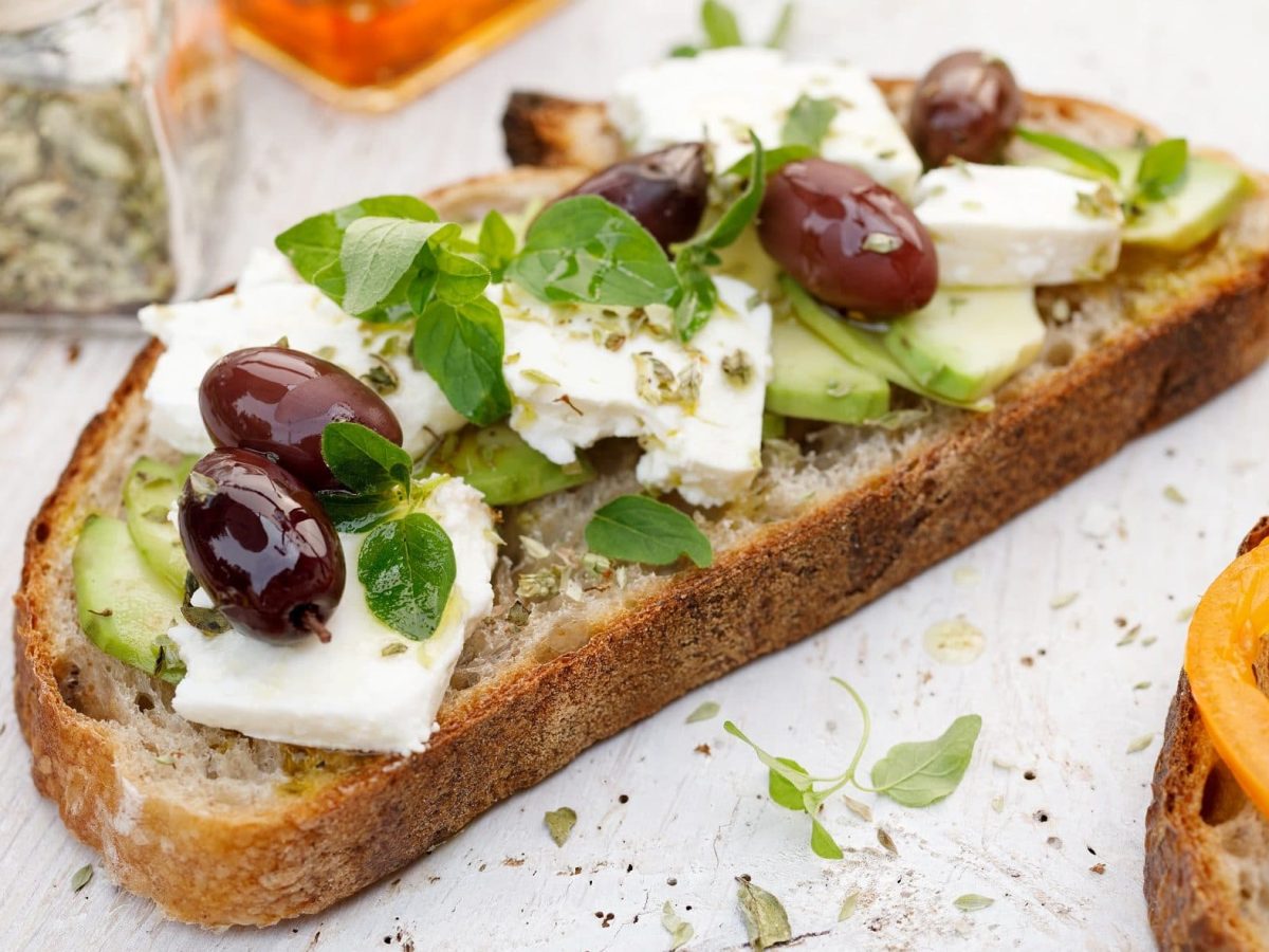 Draufsicht: Ein griechisches Sandwich mit Oliven und Feta und Oregano liegt auf einem weißen Holztisch.