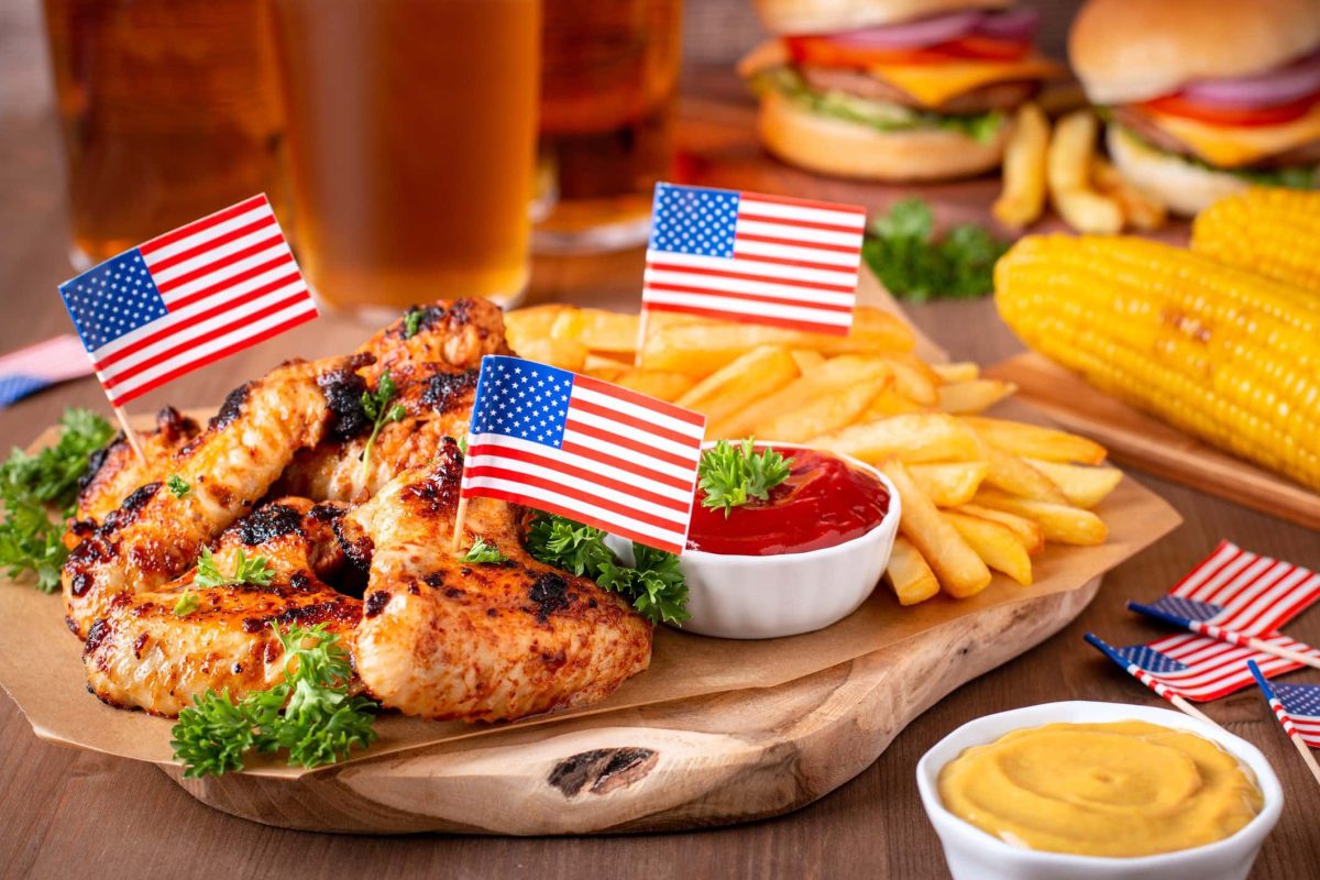Independence Day Menü amerikanische Gerichte zum 4. Juli Wings, Pimmes und weitere Gerichte mit amerikanischen Flaggen auf Brett und Holt. Ein Bier im Hintergrund. Frontalansicht.