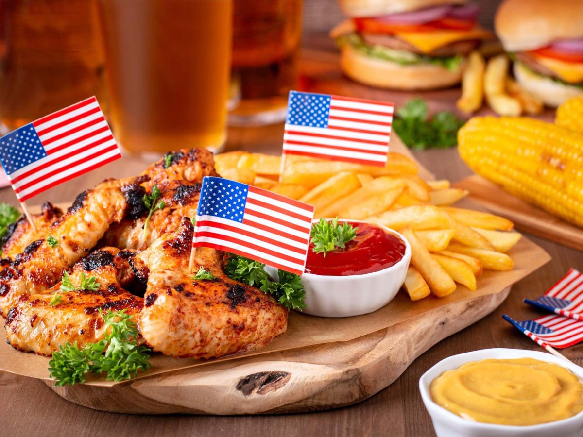 Independence Day Menü amerikanische Gerichte zum 4. Juli Wings, Pimmes und weitere Gerichte mit amerikanischen Flaggen auf Brett und Holt. Ein Bier im Hintergrund. Frontalansicht.
