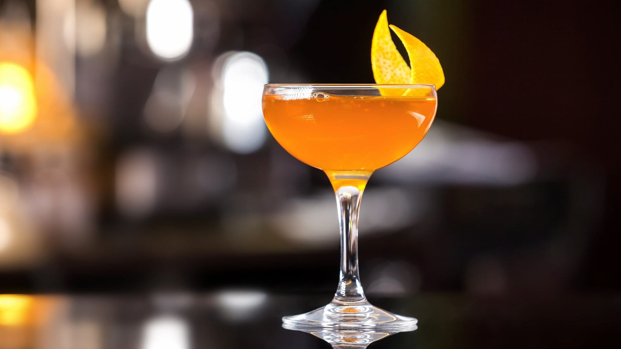 Ein Glas Italian Martini, ein orangefarbener Drink, dekoriert mit einer Zitronenzeste, in dunklem Bar-Ambiente.