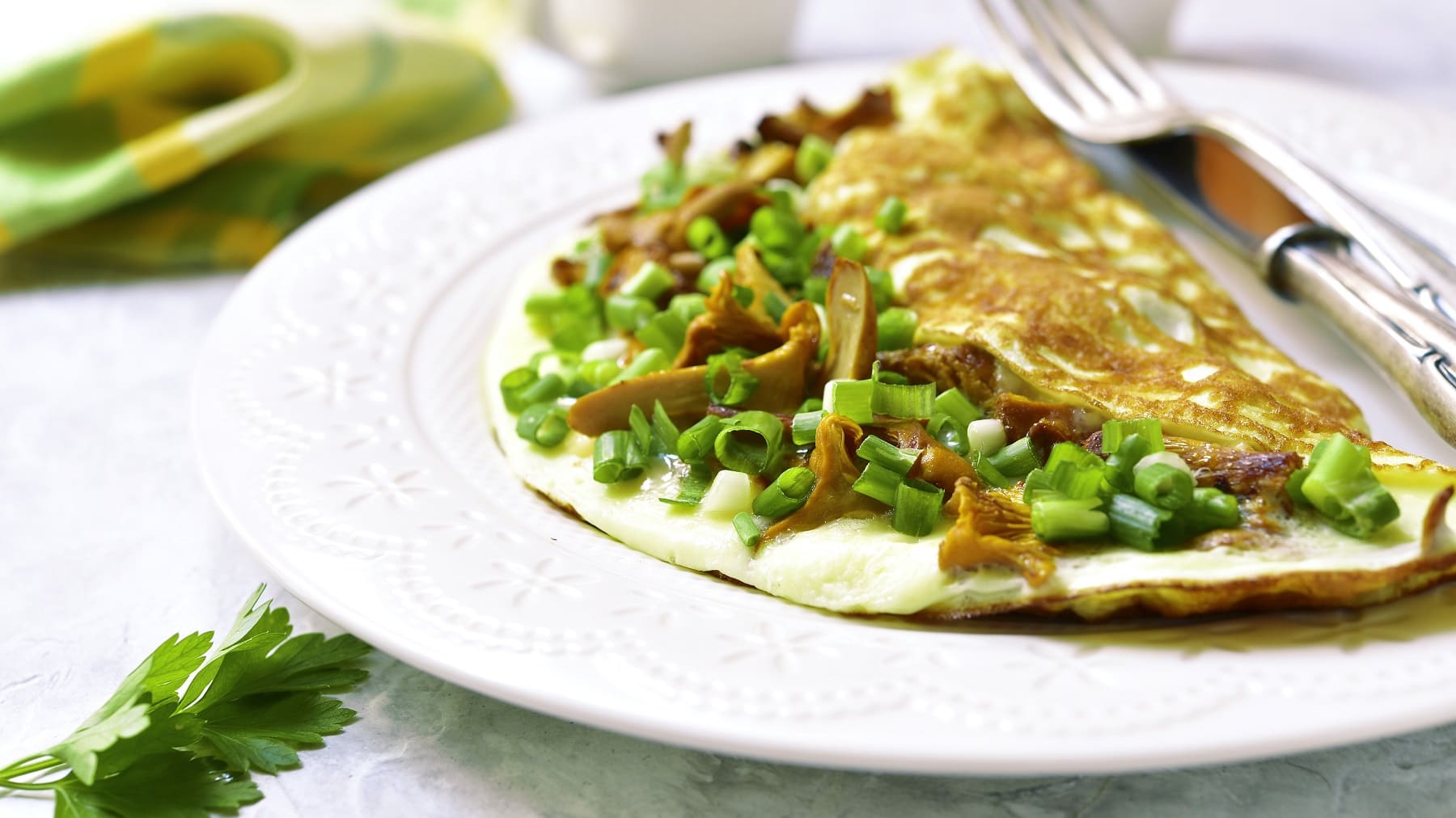 Ein weißer Teller mit dem Kräuter-Omelett und Pfifferlingen auf einem hellen Untergrund mit einem grünen Küchentuch.