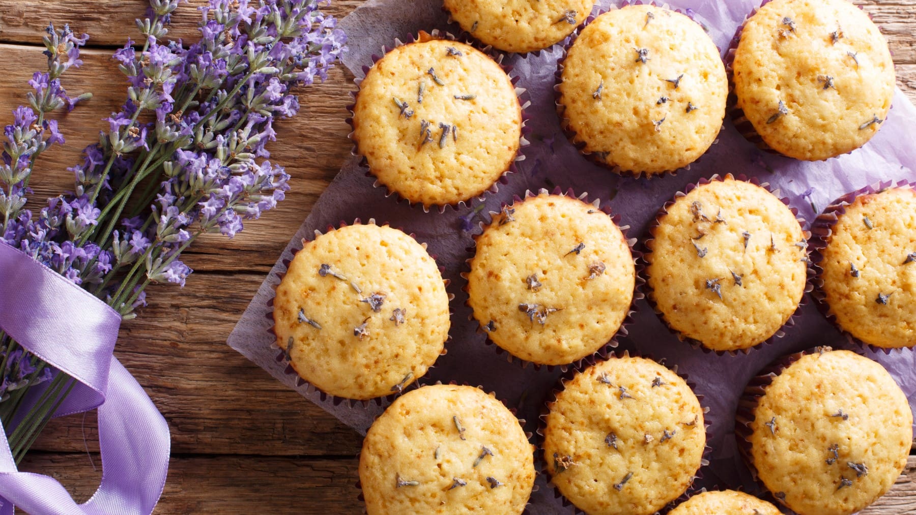 Draufsicht: Mehrere Lavendel-Muffins auf einem lila Tuch, daneben ein Bund Lavendel, Holzuntergrund.