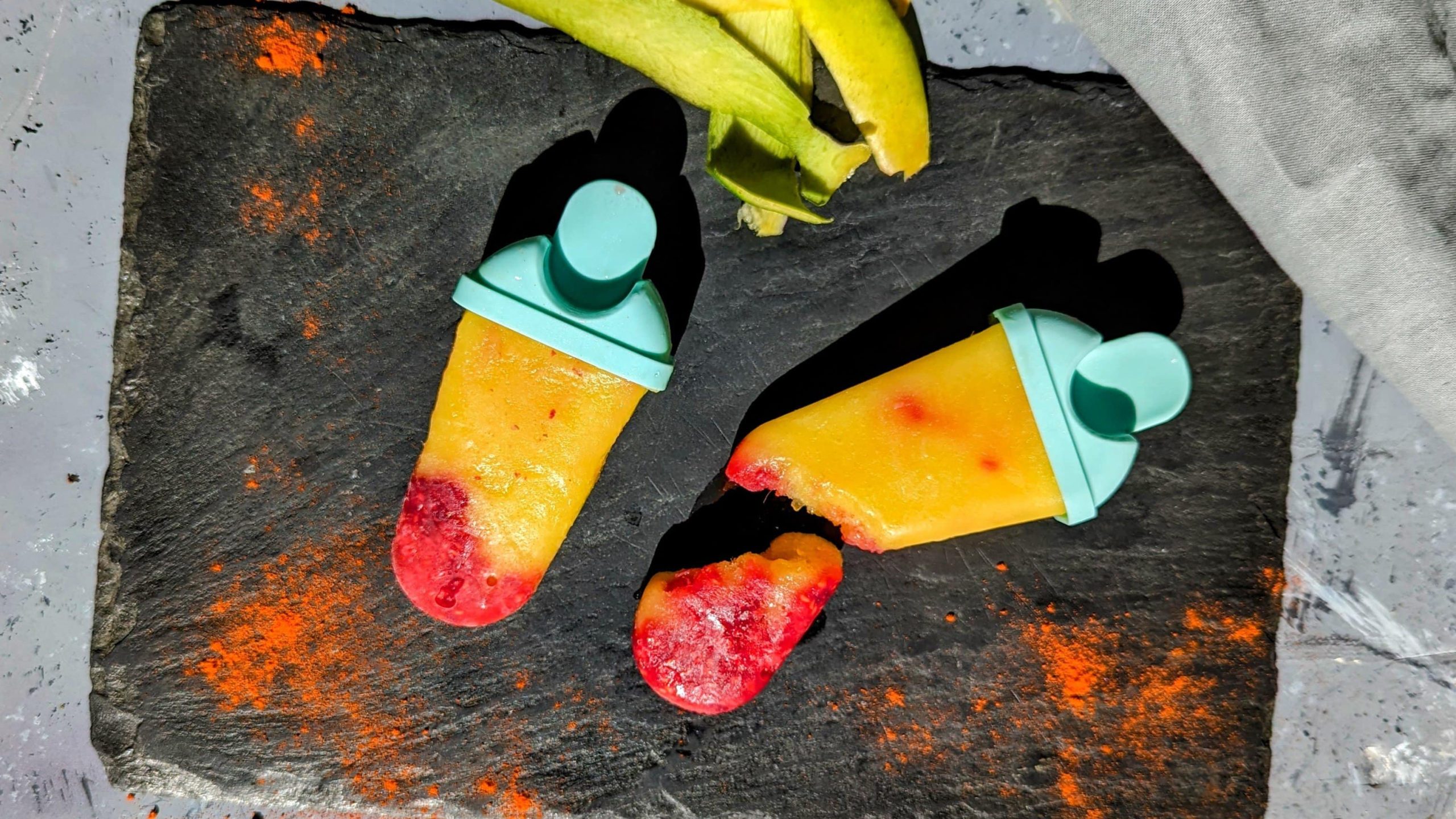 Zwei Mango-Eis mit Himbeere und Cayenne auf einer schwarzen Schieferplatte und grauem Untergrund. Daneben Mangoschalen und Cayennepfeffer.