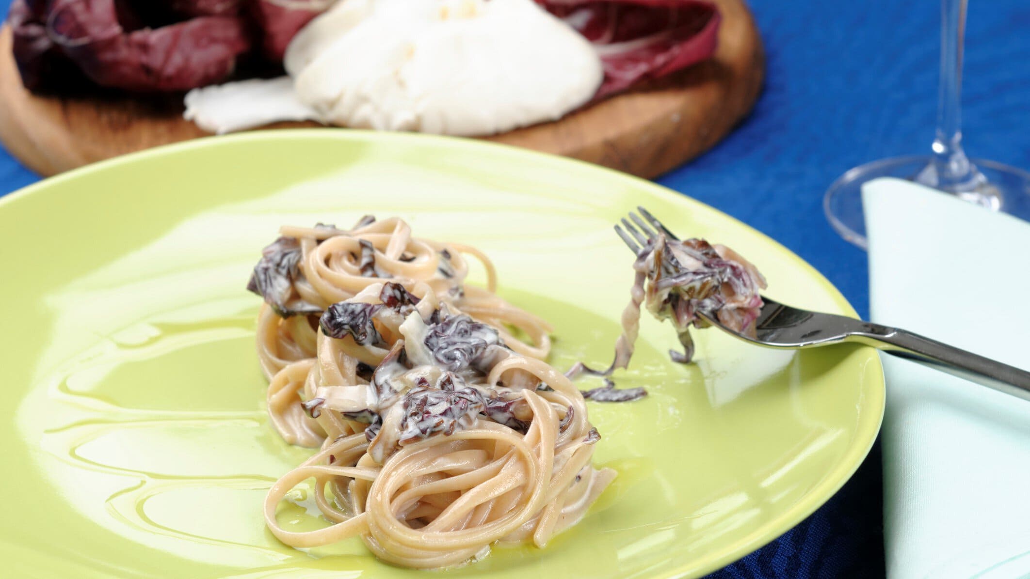 Pasta mit Radicchio und Ziegenfrischkäse auf gelbem Teller mit Gabel. Im Bild Holzbrett mit Radicchio und Ziegenfrischkäse. Draufsicht.