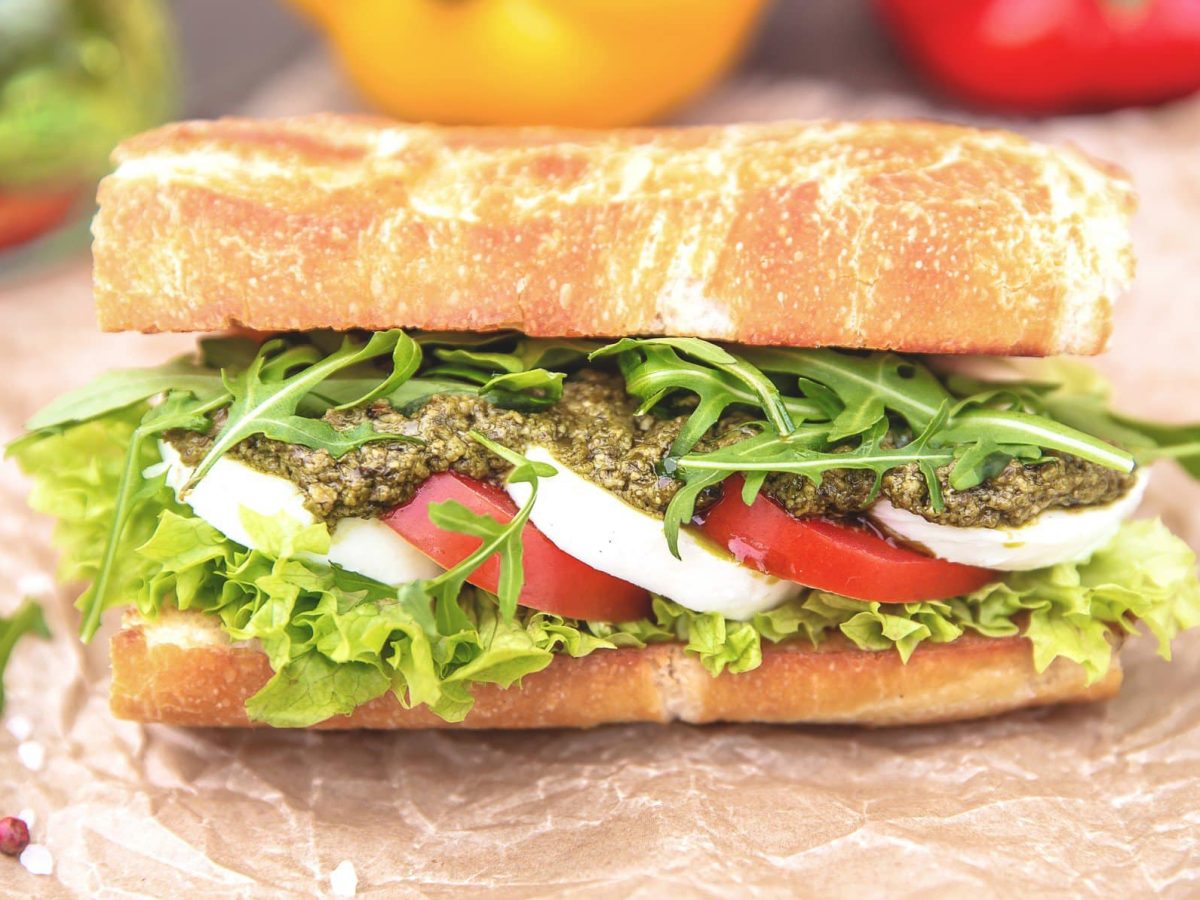 Pesto caprese Sandwich auf Papier und grauem Untergrund. Im Hintergrund Salat und buntes Gemüse. Frontalansicht.