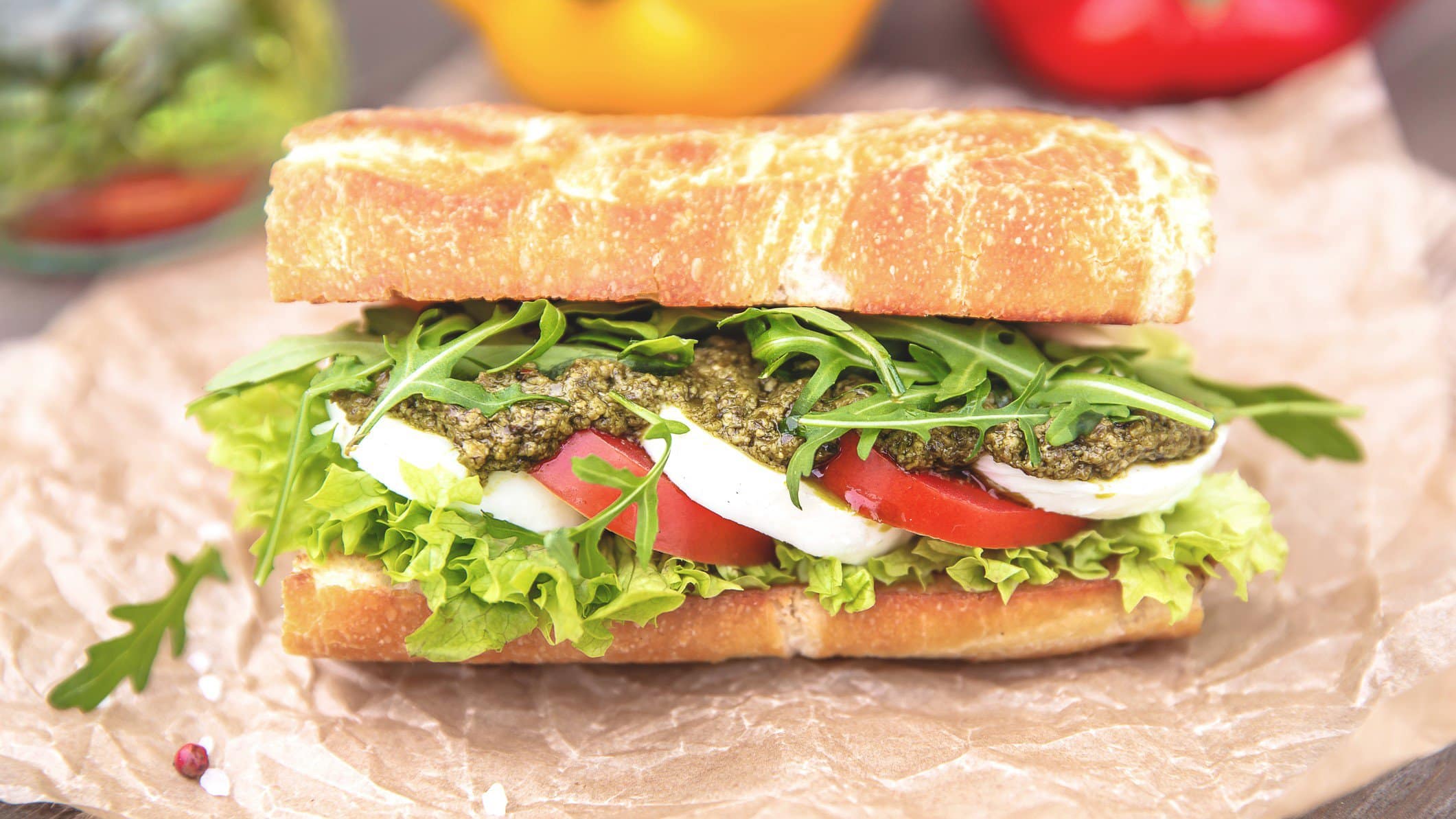 Pesto caprese Sandwich auf Papier und grauem Untergrund. Im Hintergrund Salat und buntes Gemüse. Frontalansicht.
