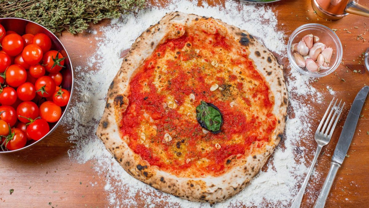 Pizza Marinara in der Draufsicht mit Knoblauch und Basilikum, daneben frische Tomaten in einer Schale, Besteck , getrockneter Oregano, eine Schale Knoblauch, eine mit Basilikum und eine Ölflasche.