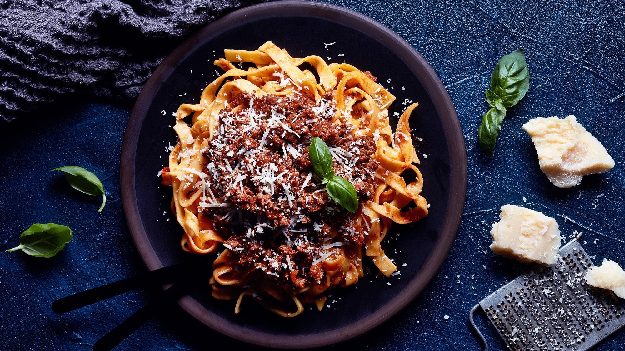 Ragù alla bolognese mit Tagliatelle, Parmesan und Basilikum auf einem Teller in der Draufsicht, daneben liegen Parmesanstücke, eine Käsereibe und Basilikumblätter.