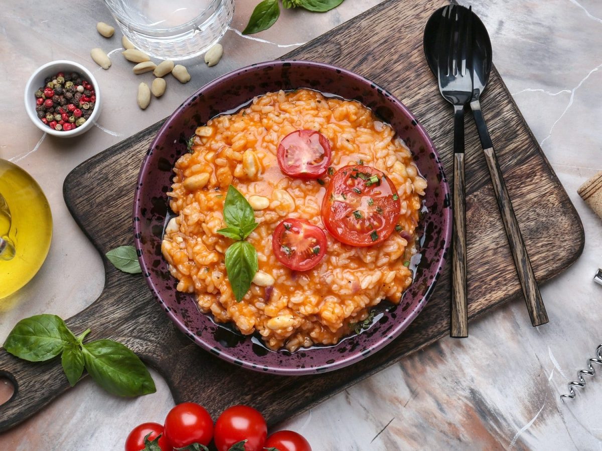Ein Teller Italienischer Tomatenreis mit Bohnen, Tomatenscheiben und Basilikum garniert, auf einem Holzbrett, daneben Löffel und Gabel sowie frische Zutaten und ein Glas Wasser.