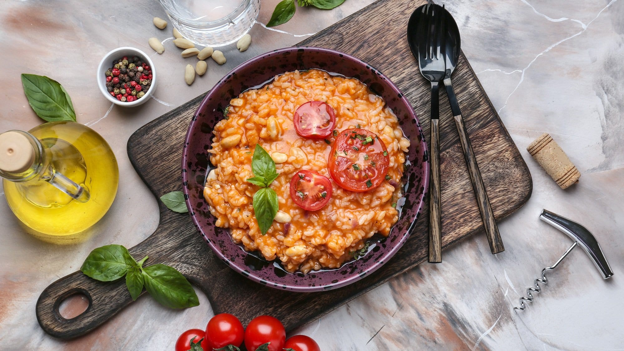 Ein Teller Italienischer Tomatenreis mit Bohnen, Tomatenscheiben und Basilikum garniert, auf einem Holzbrett, daneben Löffel und Gabel sowie frische Zutaten und ein Glas Wasser.