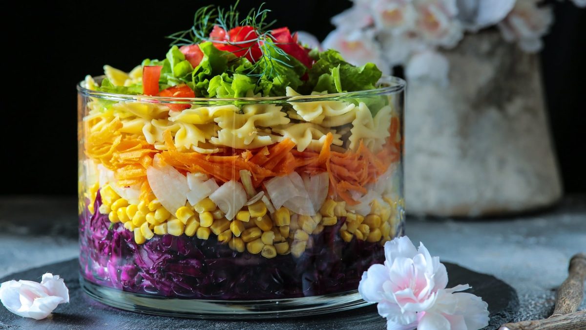 Eine große Glasschüssel mit dem geschichteten Salat auf einer schwarzen Schieferplatte mit weißen Blüten und Knospen im Hintergrund.