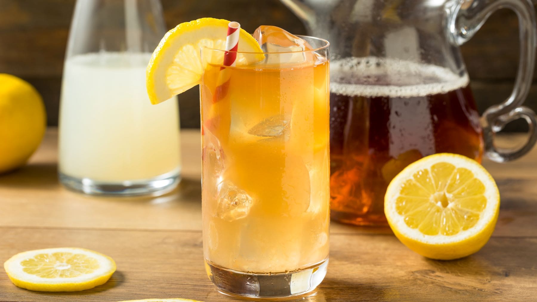 Vodka Spiked Arnold Palmer alias Jon Daly Cocktail mit Zitronenscheibe im Longdrink-Glas auf Holz. Im Bild Zitronen, Zitronenscheibe und je eine Karaffe mit Limonade und Eistee. Frontalansicht.