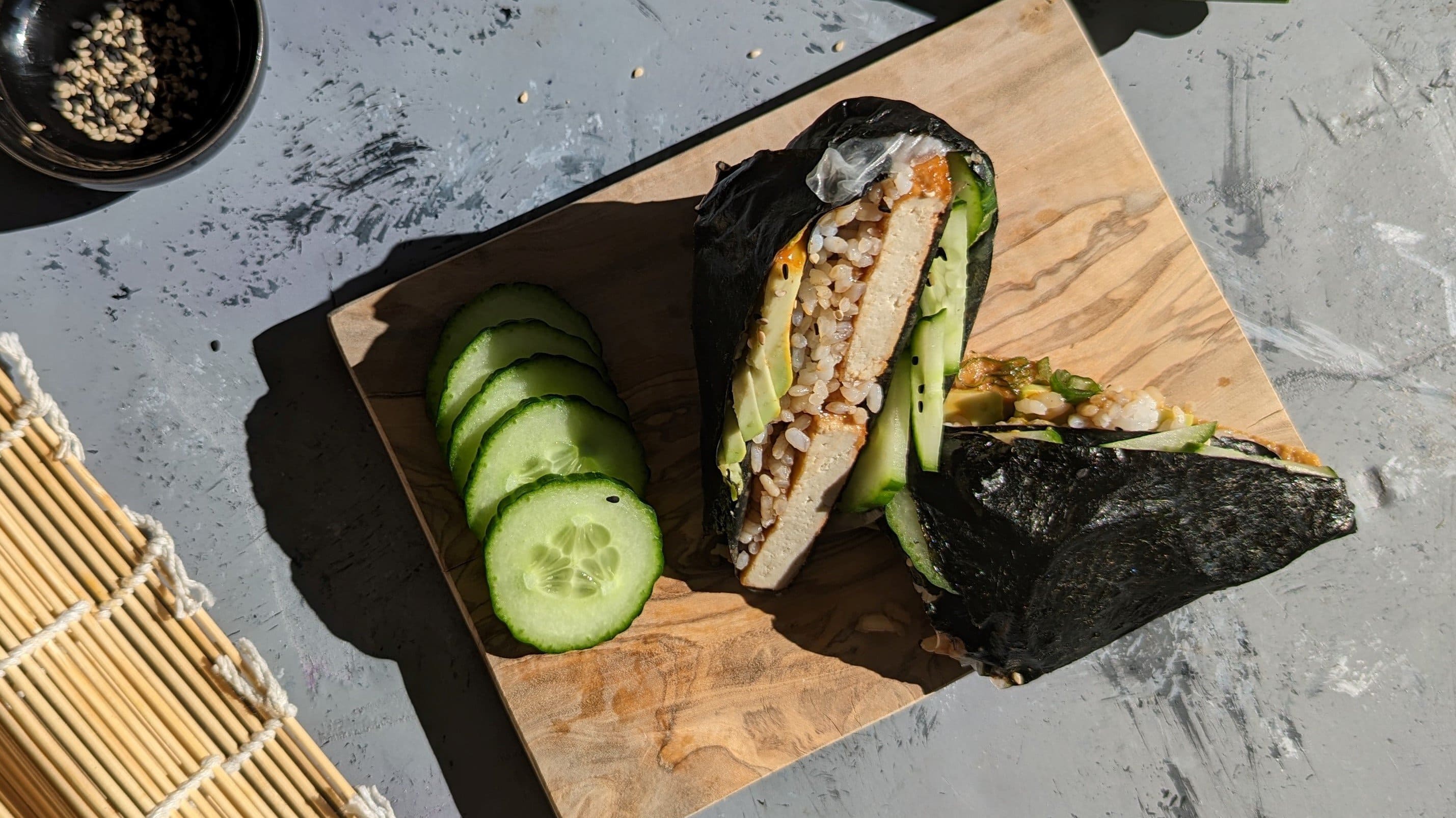 Zwei Sushi-Wraps mit einigen Gurkenscheiben auf einem kleinen Holzbrett und grauem Untergrund. Links daneben eine Sushi-Matte und ein kleines schwarzes Schälchen mit Sesam.