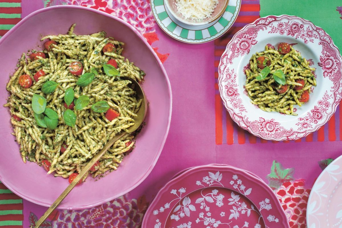 Nudeln mit Tomaten und grünem Pesto auf zwei bunt gemusterten Tellern, die auf pinkem Untergrund stehen, Draufsicht. Bild aus dem Kochbuch "Toskana".