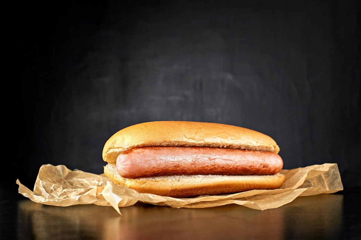 Welche Wurst für Hotdogs? Ein Frankfurter Würstchen im Hotdogbrötchen dem Betrachter zugewandt auf Backpapier vor schwarzem Hintergrund. Frontalansicht.