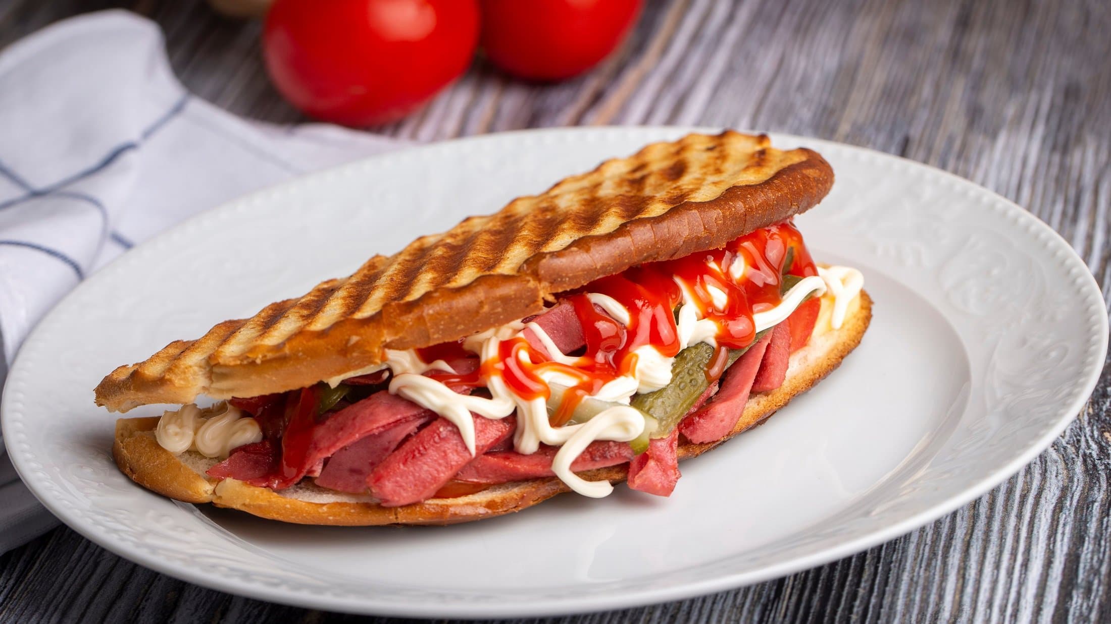Draufsicht schräg: ein türkisches Sandwich, das Ayvalık Toast mit Sucuk heißt, liegt auf einem weißen Teller. Der Untergrund ist aus Holt und darauf liegen noch zwei Tomaten und ein Tuch.