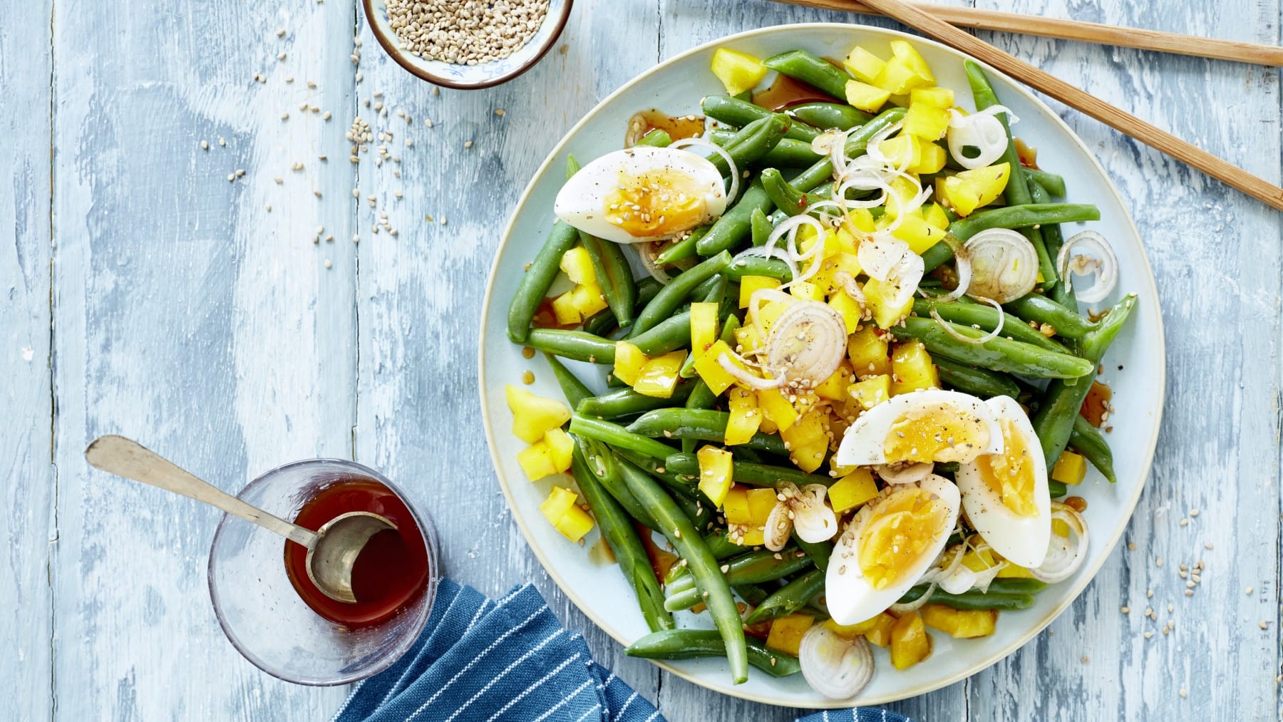 Draufsicht: Bohnensalat mit Ei und Mango auf einem Teller, daneben ein Glas mit Sojasauce unr Sesamkörner auf blauem Holzuntergrund.