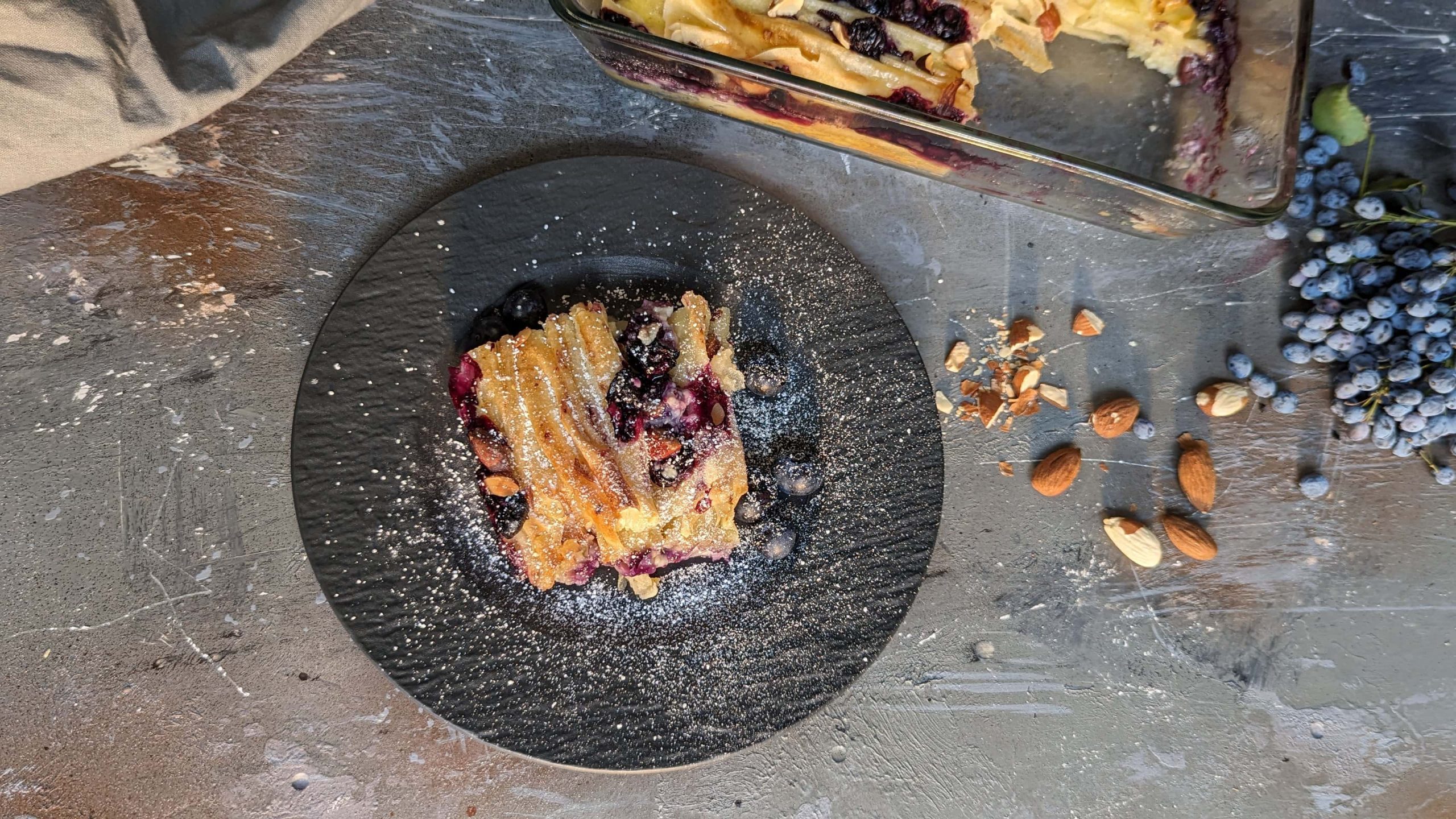 Ein schwarzer Teller mit einem Stück vom Crinkle Cake auf grauem Steinuntergrund. Daneben die Glas-Auflaufform mit dem Rest. Alles von oben fotografiert.