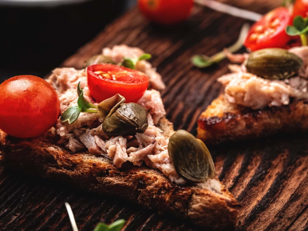 Geröstetes Knoblauch-Sandwich mit Thunfisch, Kapern und Tomaten auf Holzbrett mit Kressen. Im Bild Kapern. Draufsicht.