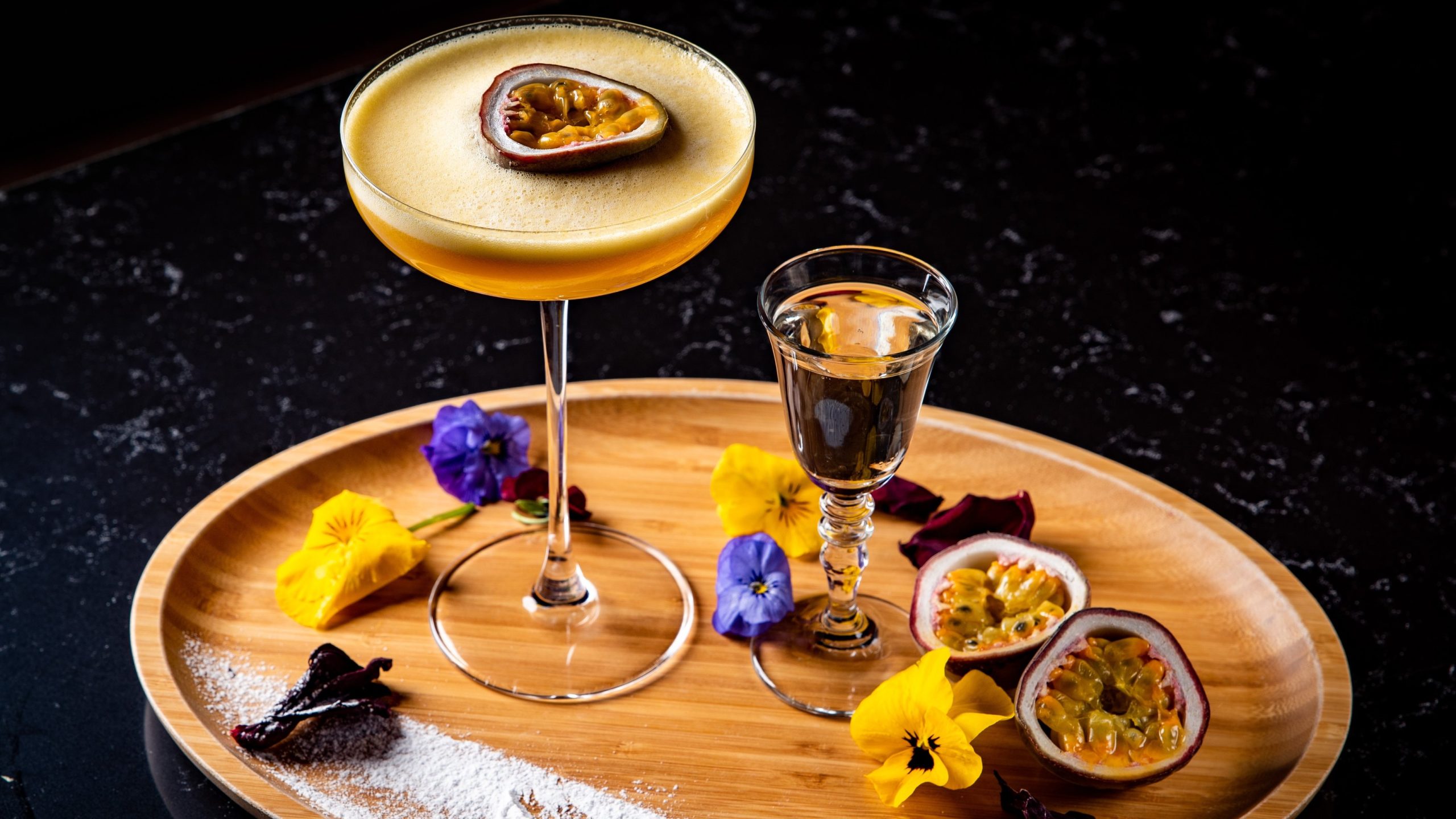 Ein Glas mit dem Pornstar Martini und einem Shotglas mit Champagner auf einem hellen Holzbrett mit violetten und gelben Blüten. Alles auf einem dunklen Tisch.