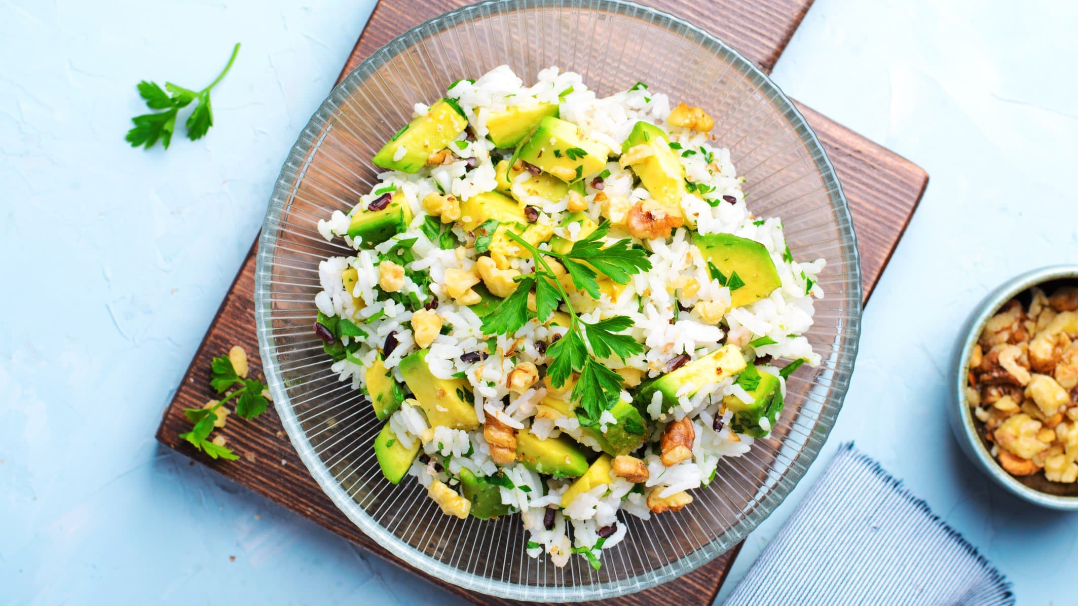 Draufsicht: Ein Teller Reissalat mit Avocado und Nüssen auf einem hellblauen Untergrund.