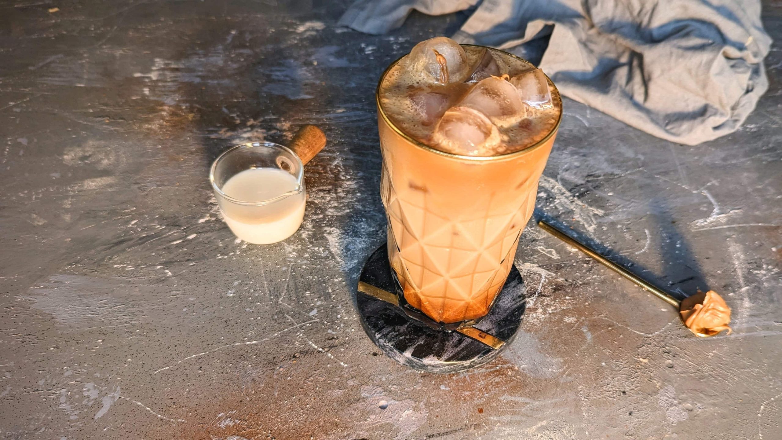 Ein Glas mit Caramel-Coffee auf grauem Untergrund und schwarzem Coaster. Außerdem ein Löffel mit Karamell und ein Gläschen mit Hafermilch.