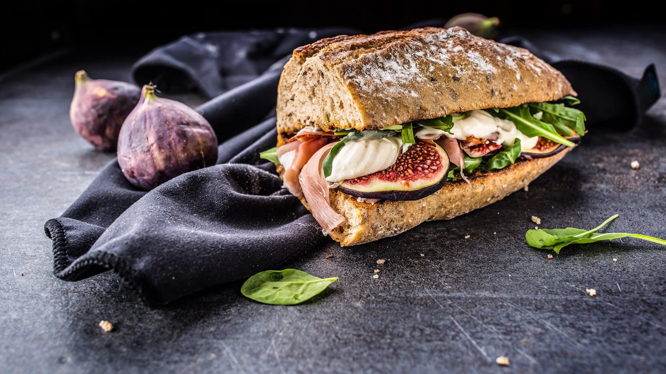 Frontal: Ein Schinken-Feigen-Sandwich mit Mozzarella auf einem Tuch, das auf dunklem Untergrund liegt. Drum herum liegen frische Feigen und Salatblätter.