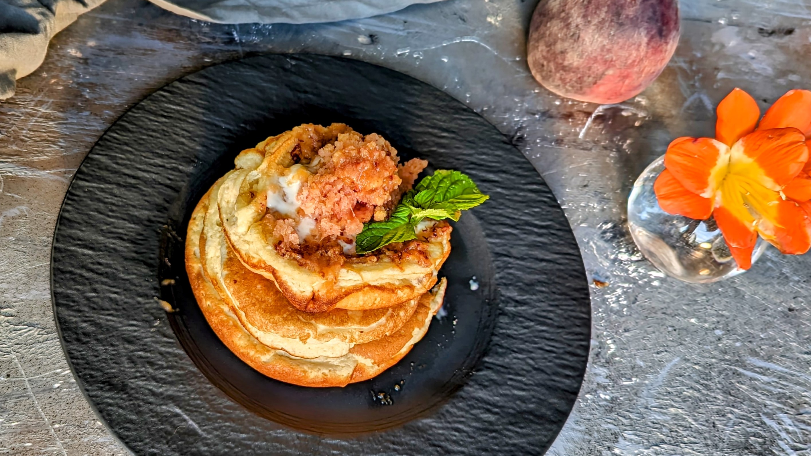 Ein schwarzer Teller mit Pancakes und geriebenem, gefrorenem Pfirsich auf grauem Steinuntergrund. Daneben eine orangene Blüte, ein Pfirsich sowie eine graue Tischdecke.