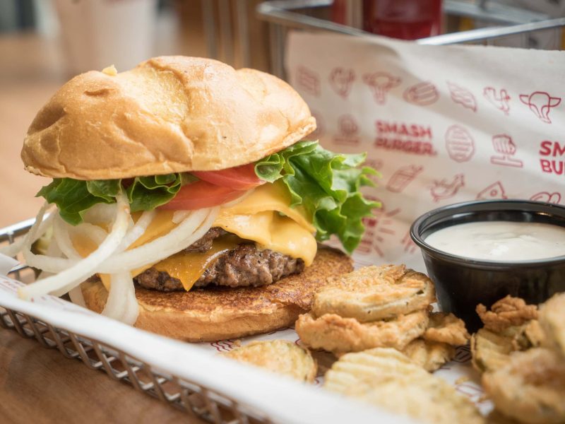 Was ist ein Smachburger? Unterschied zwischen Smashburger und normalem Burger. Smashburger mit frittiertem Gemüse und Dip auf weißer Servierplatte. Im Hintergrund eine Papier der Marke Smashburger. Frontalansicht.