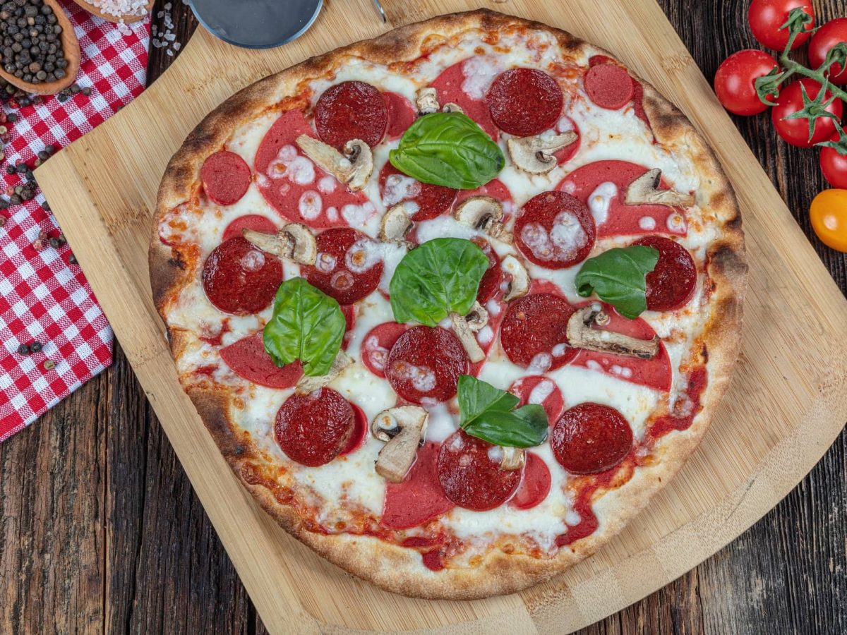 Draufsicht: dunkler Untergrund, rot-weiß kariertes Tuch, Gewürze, frische Tomaten und Basilikumblätter. Mittendrin liegt eine runde Sucuk-Pizza.
