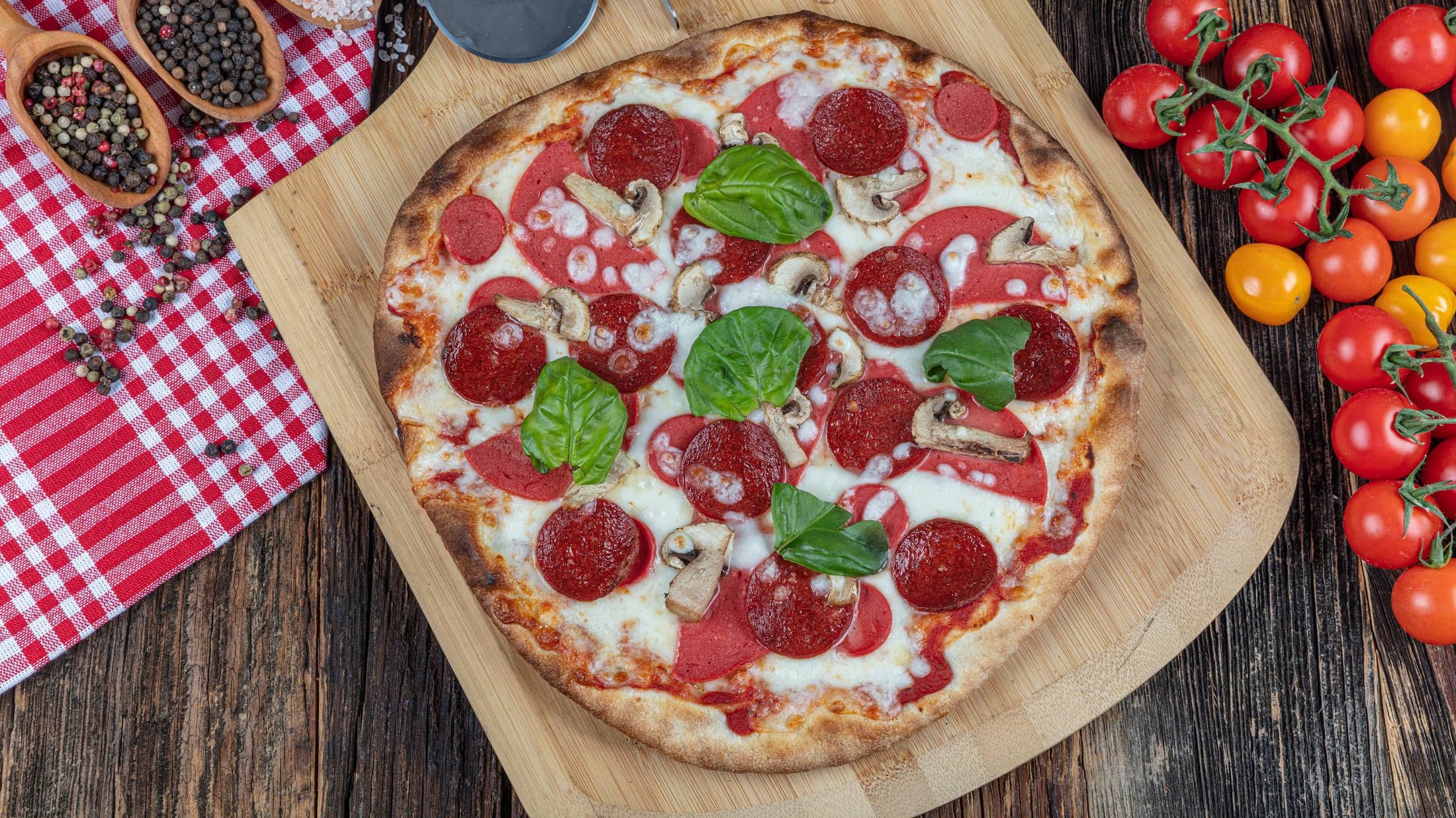 Draufsicht: dunkler Untergrund, rot-weiß kariertes Tuch, Gewürze, frische Tomaten und Basilikumblätter. Mittendrin liegt eine runde Sucuk-Pizza.