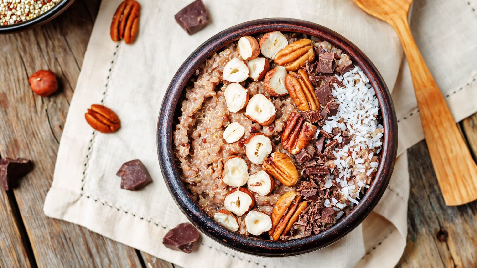 Eine Schüssel süßer Quinoa mit Nüssen, Schokolade und Kokosraspeln in der Draufsicht, daneben ein Holzlöffel und Zutaten.