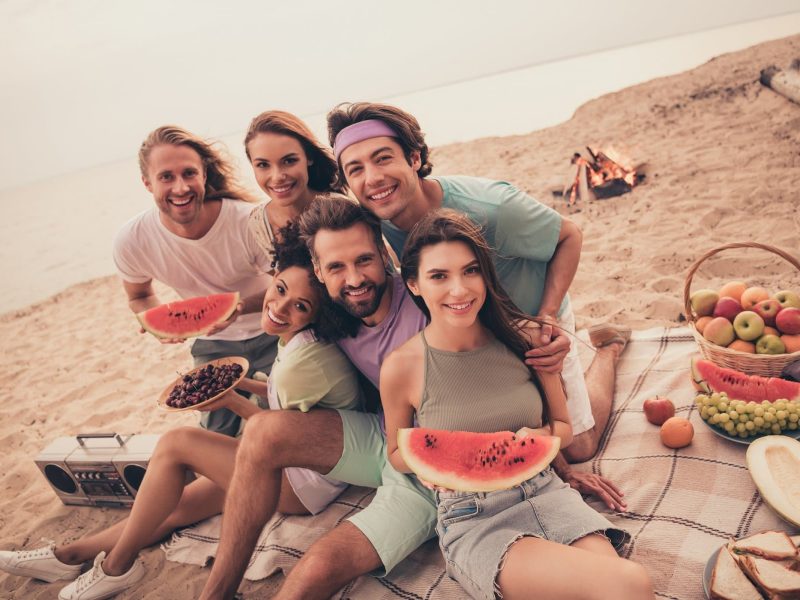 Strand, Hintergrund Meer. Eine Gruppe fröhlicher jungen Menschen mit Wassermelonen in den Händen schauen frontal in die Kamera.
