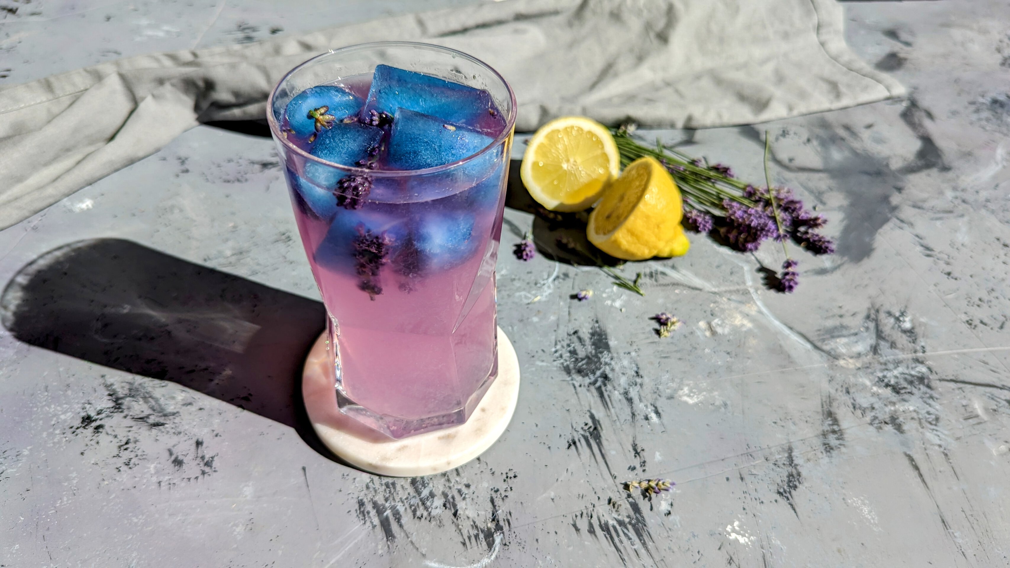Ein Glas mit der Zitronenlimonade und den blauen Eiswürfeln auf einem Steintisch. Die Limonade hat sich schon leicht pink verfärbt. Im Hintergrund Zitrone und Lavendel.