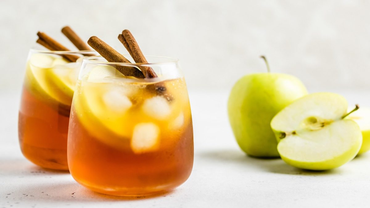 Zwei Gläser Apple Cider Spritz mit Eiswürfeln, Apfelscheiben und Zimtstangen garniert, daneben liegen ein ganzer und ein angeschnittener Apfel.