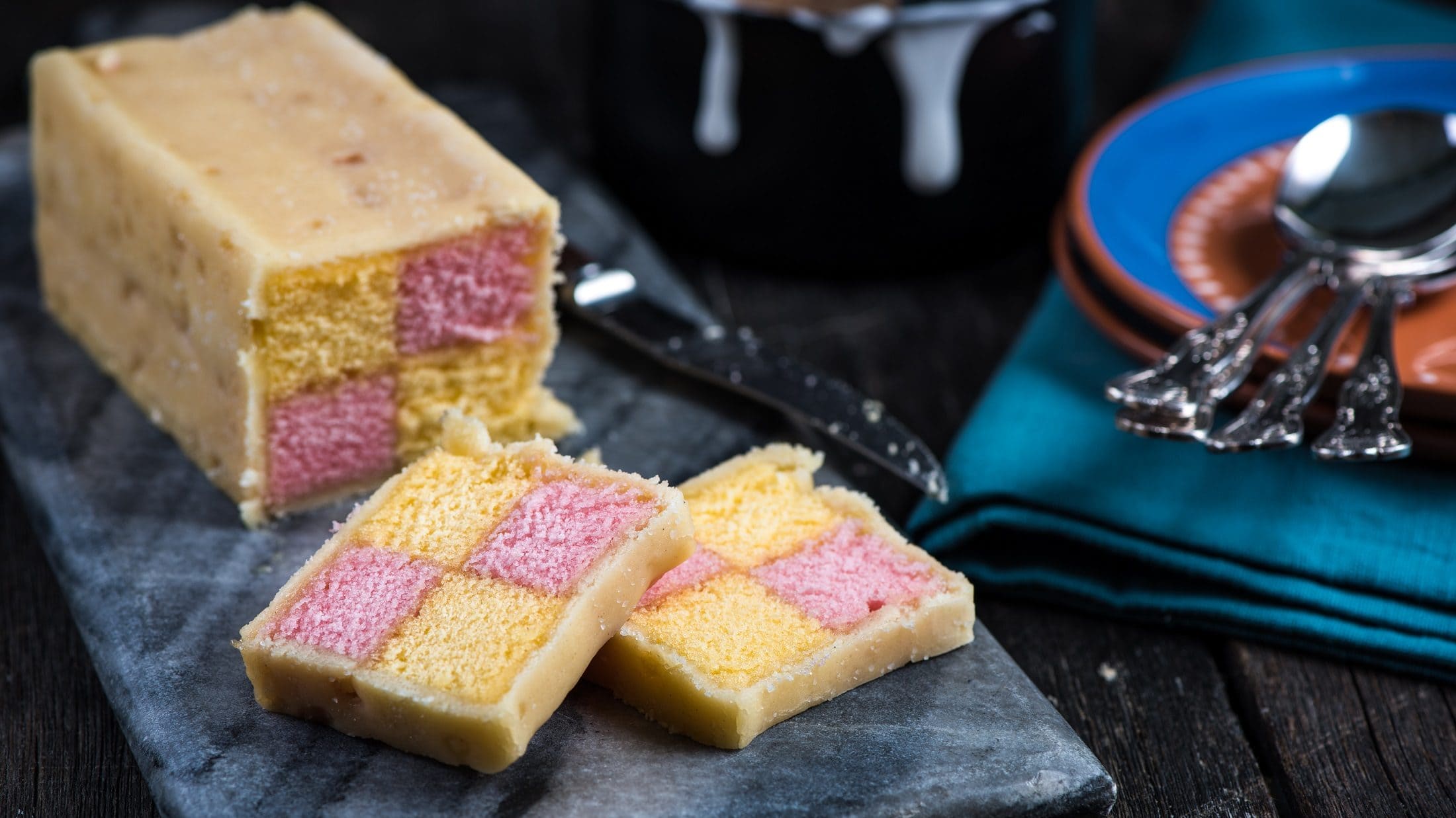 Draufsichtschräg: ein angeschnittener Battenberg Cake auf einem dunklen Brettchen auf dunklem Untergrund. Der Kuchen hat innen zwei verschiedene Farben, die Karo-Förmig angeordnet sind. Es handelt sich um die Farben gelb und rosa.