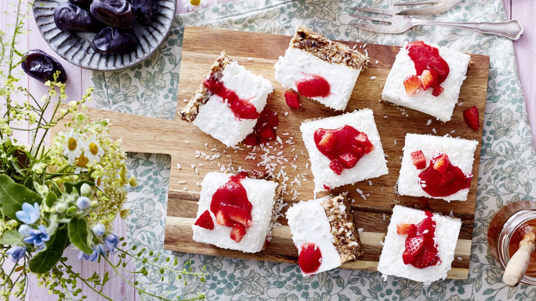 8 Stücke Dattel-Joghurt-Cheesecake auf einem hölzernen Schneidebrett auf einem Geschirrtuch. Rechts unten Honig, links unten Blumen. Links oben eine kleine Schüssel mit Datteln und rechts oben zwei Dessertgabeln.