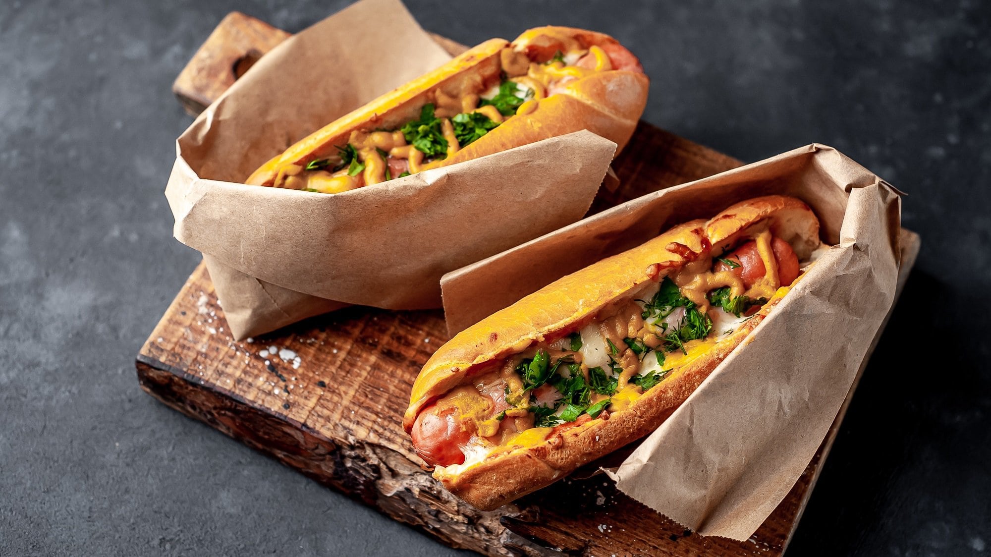 Zwei Hot Dogs mit selbst gemachter Hot-Dog-Sauce, Käse und Petersilie, eingewickelt in Papier, auf einem Holzbrett.