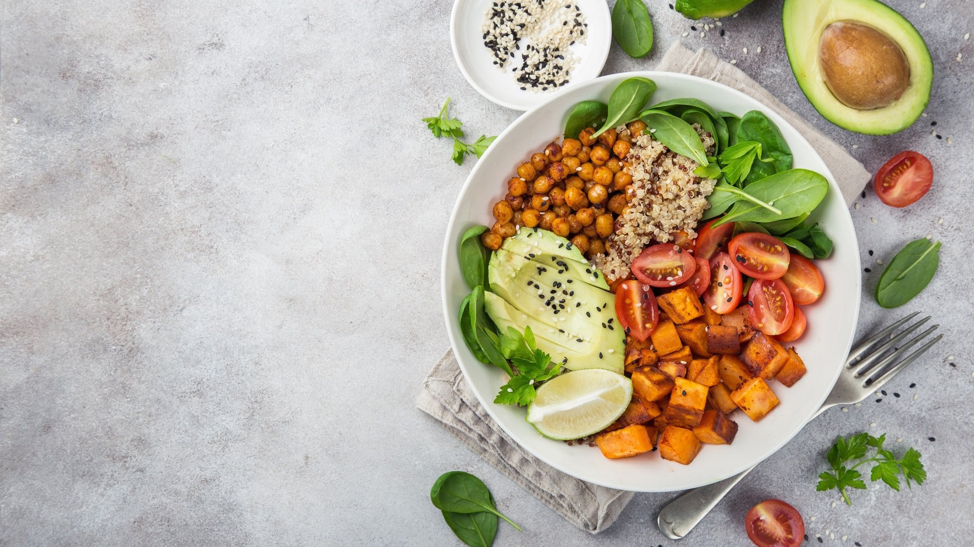 Eine Kürbis-Quinoa-Bowl in der Draufsicht mit Babysoinat, Kichererbsen und Avocado sowie einer Spalte Limette, drumherum frische Zutaten.