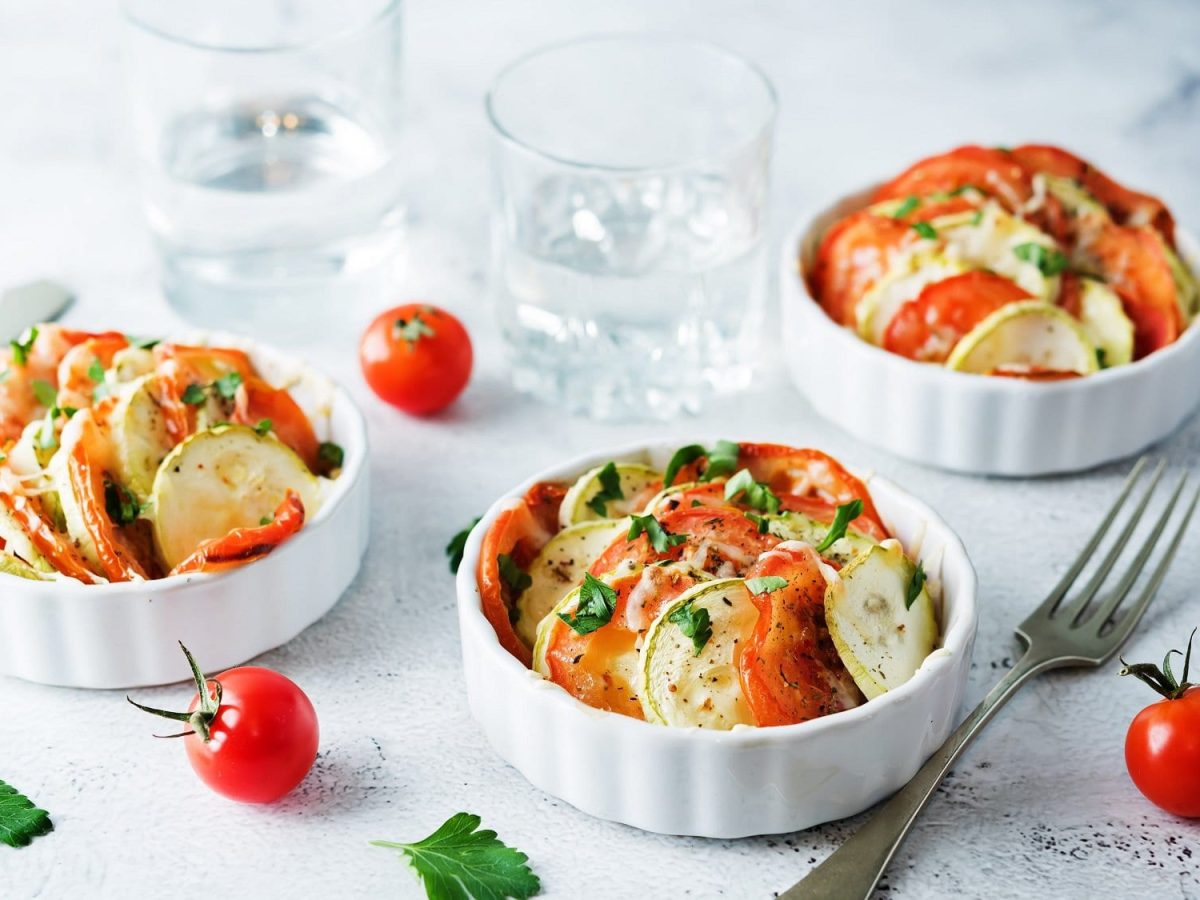 Mini-Gemüse-Gratins mit Zucchini und Tomate in kleinen runden weißen Auflaufformen, drumherum liegen eine Gabel, Tomaten und Petersilienblätter.