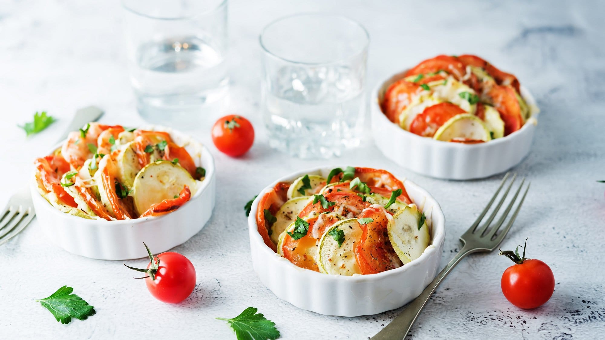 Mini-Gemüse-Gratins mit Zucchini und Tomate in kleinen runden weißen Auflaufformen, drumherum liegen eine Gabel, Tomaten und Petersilienblätter.