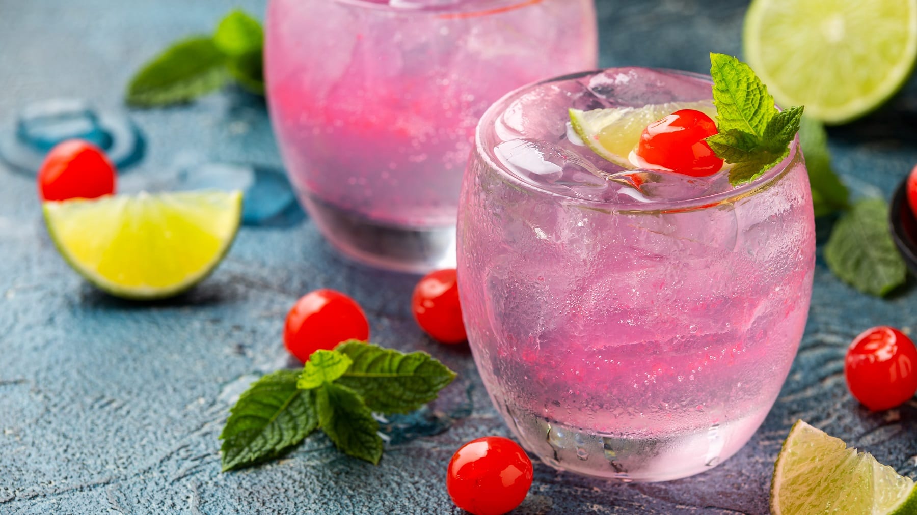 Zwei Gläser mit dem Pink Cherry Gin auf dunkelblauem Tisch mit Limettenspalten und Maraschino-Kirschen dekoriert. Alles seitlich fotografiert.