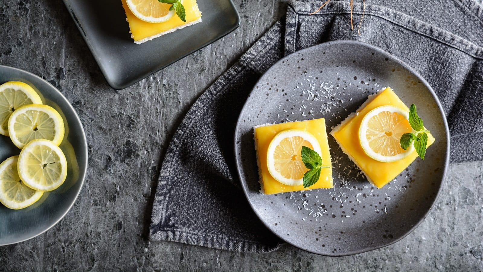 Ein grauer Teller mit zwei Kokos-Zitronen-Schnitten auf einem grauen Tisch.